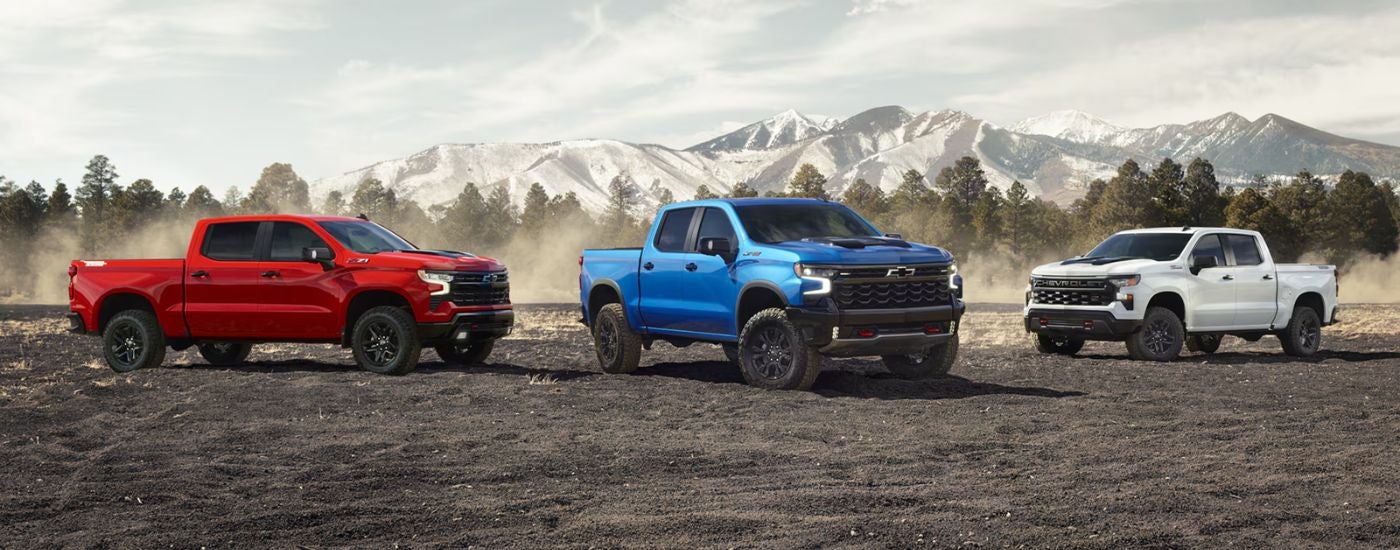 A trio of 2026 Chevy Silverado 1500s in red, white and blue in front of a mountain.